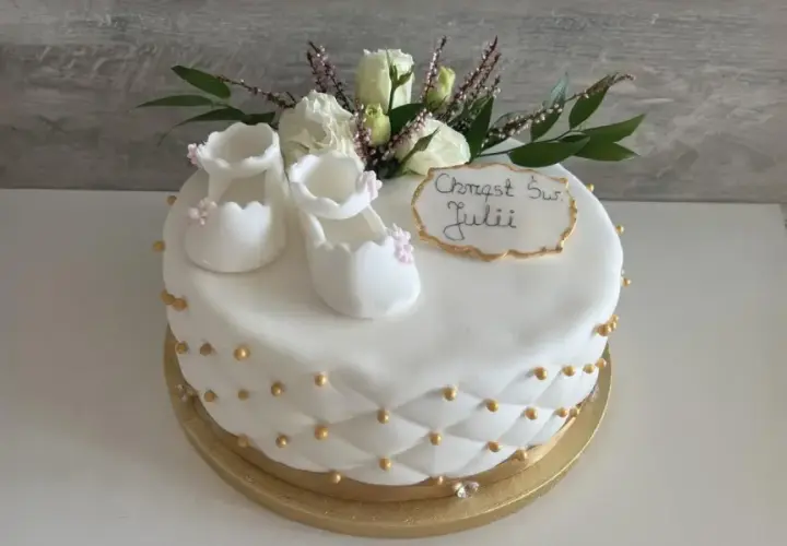 Delikatny tort na chrzest dla Julii, ozdobiony białymi bucikami, kwiatami i złotymi perełkami.