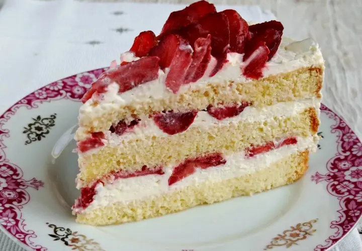 Pyszny tort z bitą śmietaną i truskawkami, przełożony kremem i owocami, na ozdobnym talerzyku.